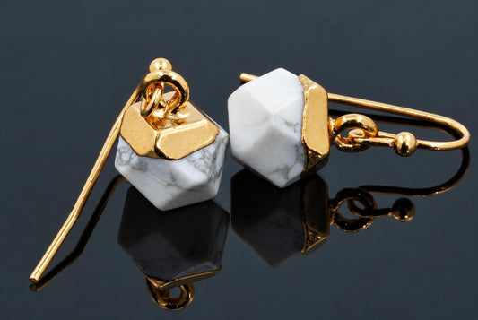 A pair of gold-toned lever-back earrings with white and grey veined faceted cube stones on a dark reflective surface.