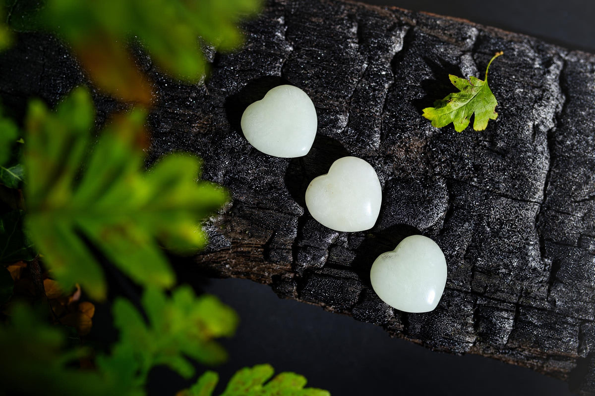 Pale green polished heart-shaped stones form a diagonal row on a dark textured surface among green leaves.