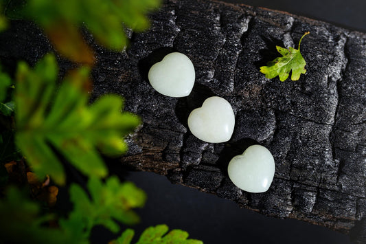 Pale green polished heart-shaped stones form a diagonal row on a dark textured surface among green leaves.