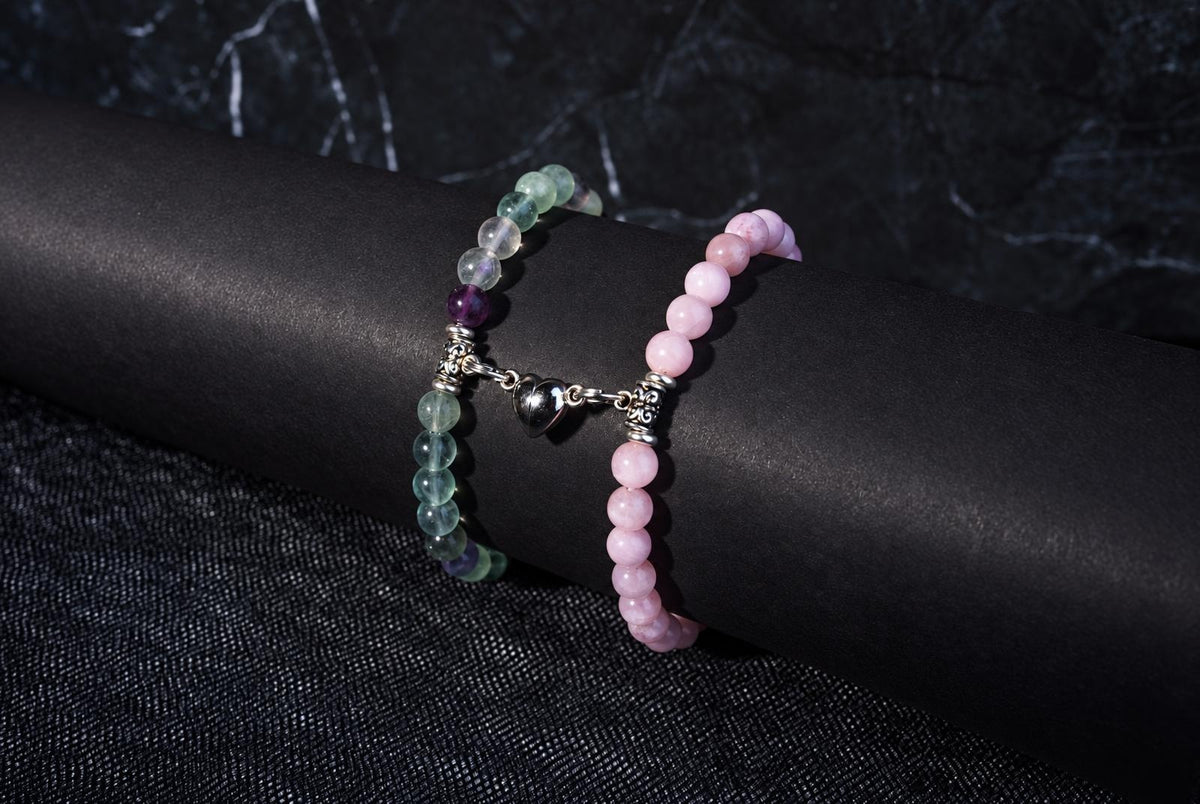 Two beaded bracelets on a dark surface: one with pink beads, the other with green and purple beads linked by a silver heart charm.