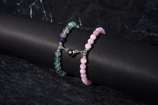 Two beaded bracelets on a dark surface: one with pink beads, the other with green and purple beads linked by a silver heart charm.