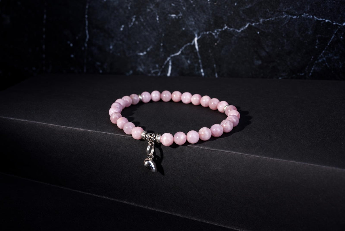 Kunzite bracelet with pale pink round beads, ornate silver spacers and a hanging heart charm against a dark background.