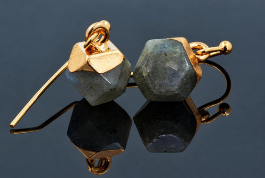 A pair of gold-toned hook earrings with faceted, blue-grey Labradorite stones set in geometric frames on a dark reflective surface.
