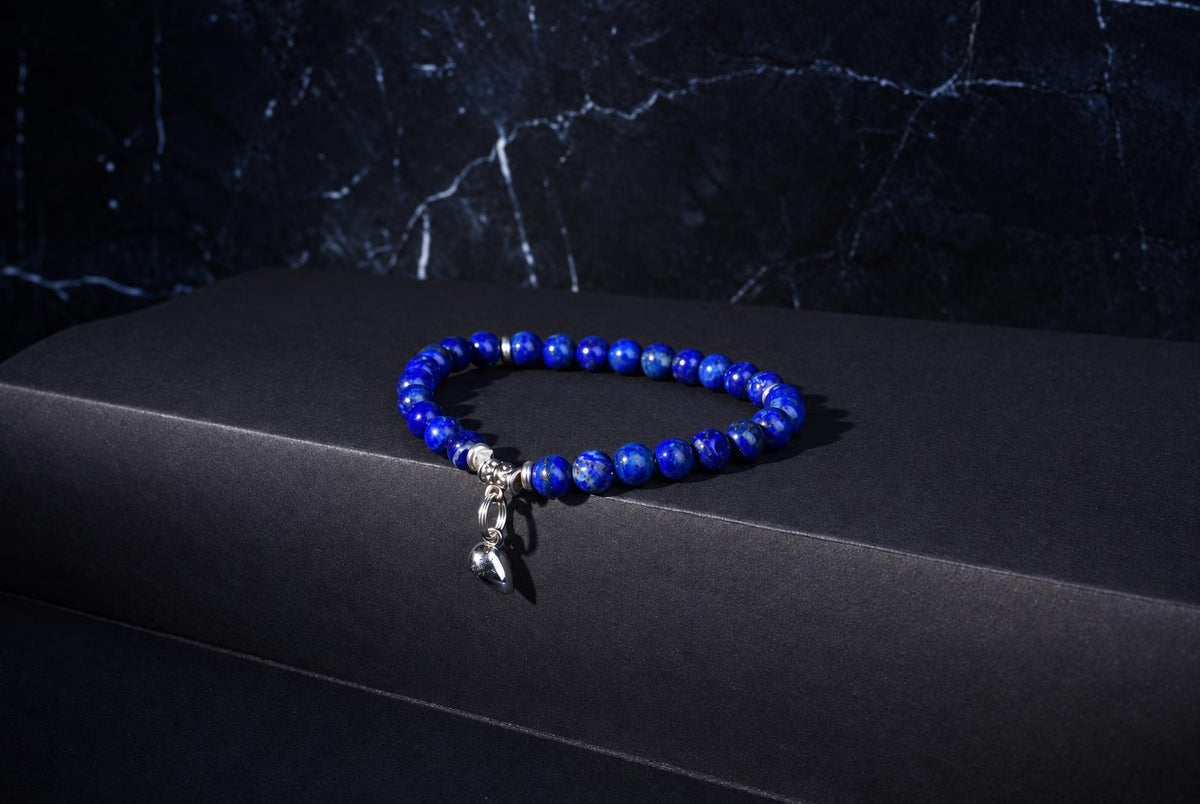 A blue beaded bracelet with silver accents and a small heart-shaped charm rests on a dark surface.