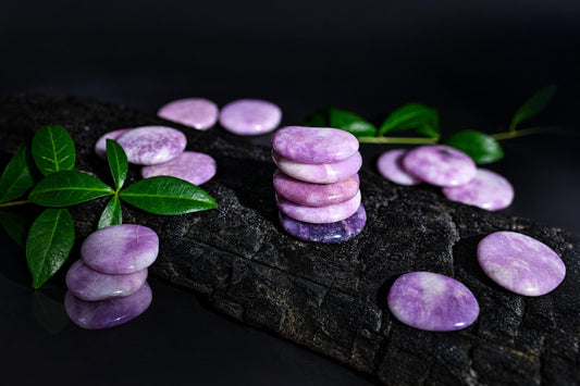 Polished purple oval stones are stacked and scattered on a dark textured surface among green leaves.