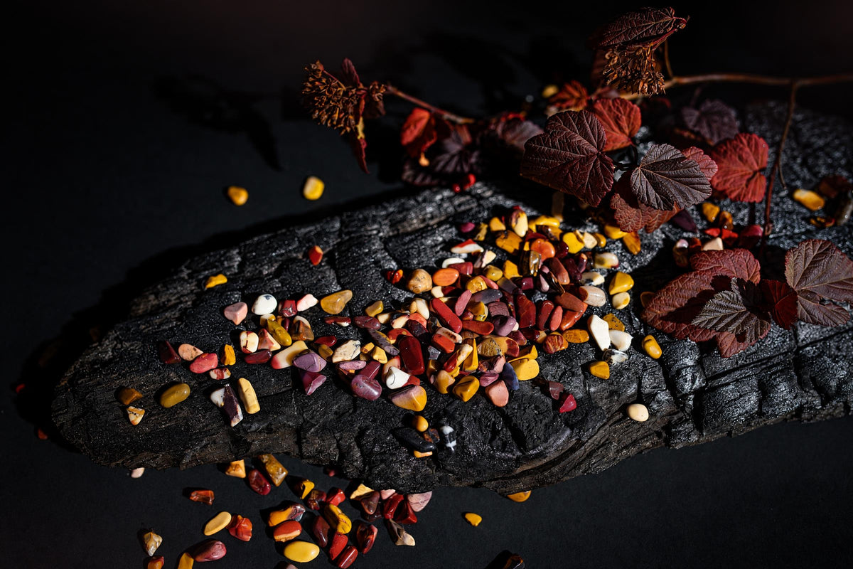 Small polished red, yellow and cream stones are scattered on a dark textured surface beside reddish leaves.