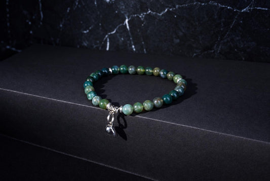 Moss agate bracelet with green round beads, ornate silver spacers and a dangling heart charm against a dark background.