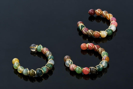 Open circular rings of colourful polished beads wrapped in gold-coloured wire rest on a dark reflective surface.