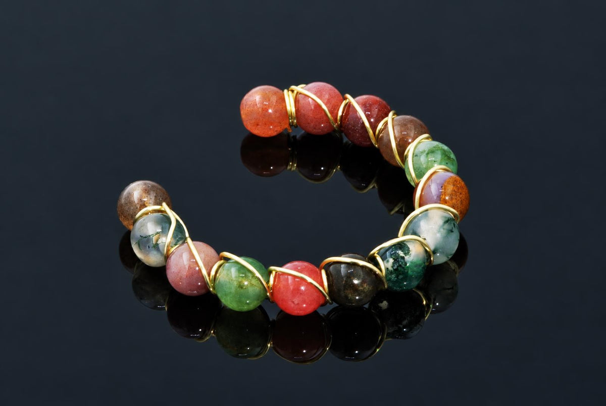 An open circular ring of colourful polished beads wrapped in gold-coloured wire rests on a dark reflective surface.