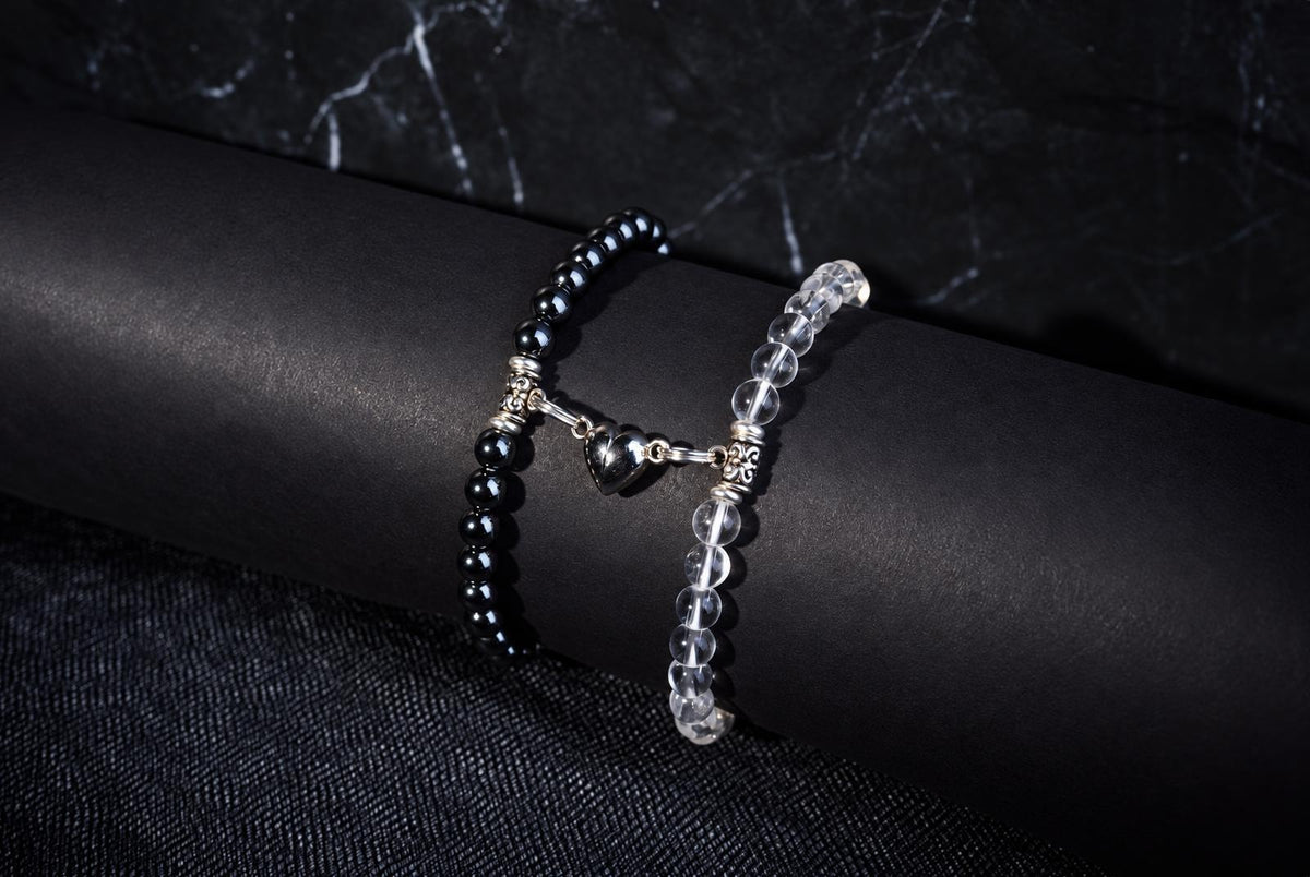 Two beaded bracelets resting on a black cylindrical display stand. One bracelet features opaque black beads and the other features clear, transparent beads. Both are connected by a silver metal charm shaped like a heart.
