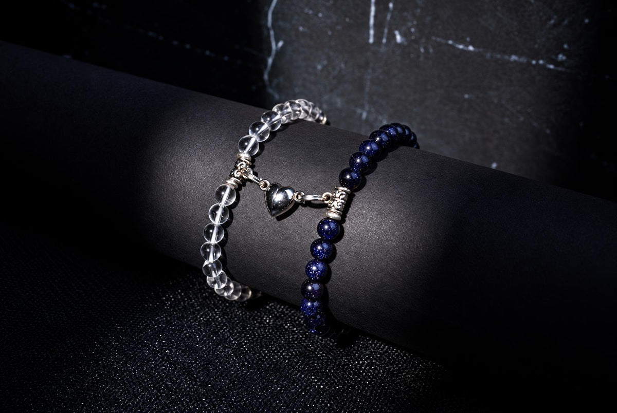 Two beaded bracelets on a dark cylindrical surface: one with clear beads and a silver heart charm, the other with deep blue beads.