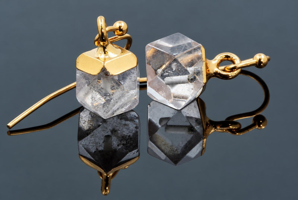 A pair of gold-coloured hook earrings with faceted, transparent cube-shaped stones on a dark reflective surface.