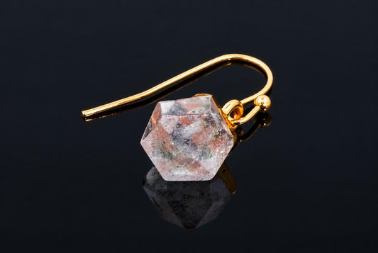 A clear faceted cube earring with pink and dark flecks and a gold-coloured hook rests on a dark reflective surface.