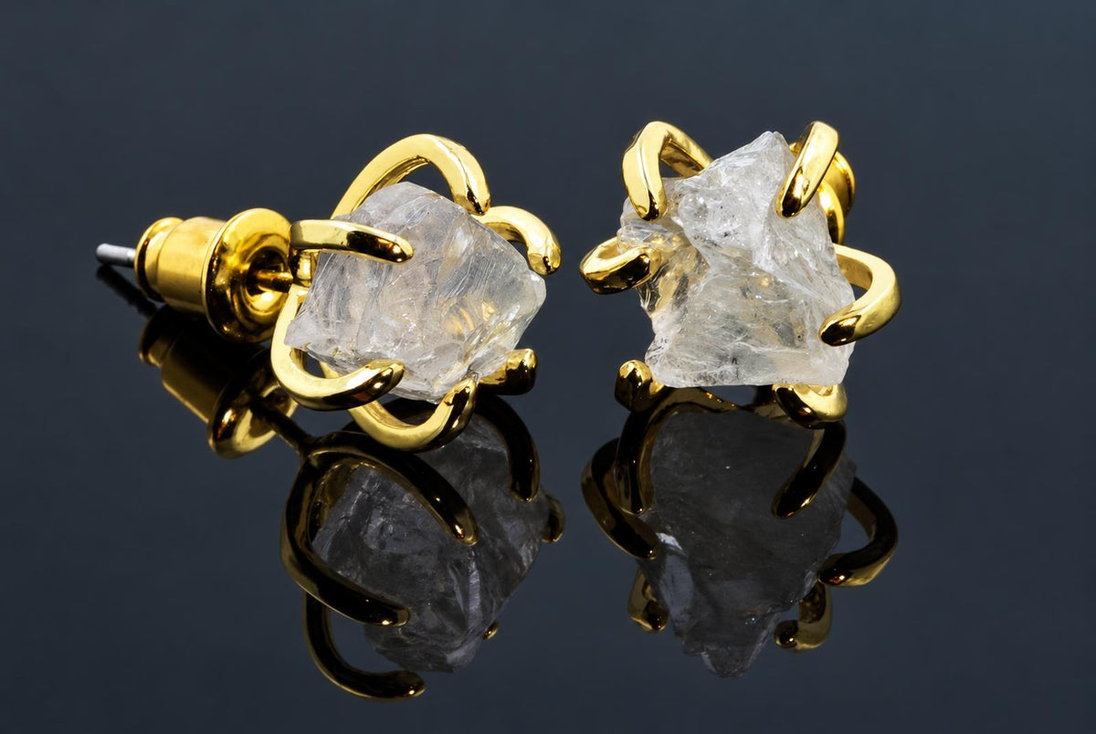 Clear rough crystal stud earrings in curved gold-coloured settings rest on a dark reflective surface.