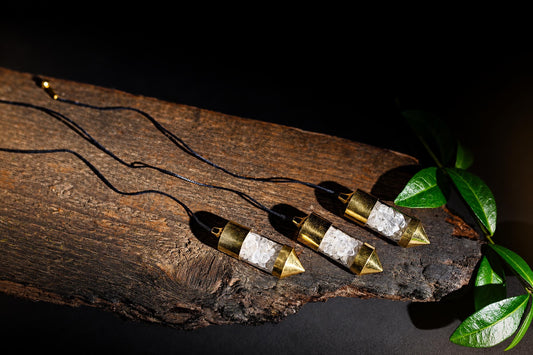 Pendulums with clear stone chips in transparent tubes and gold-coloured ends lie on wood with black cords and green leaves.