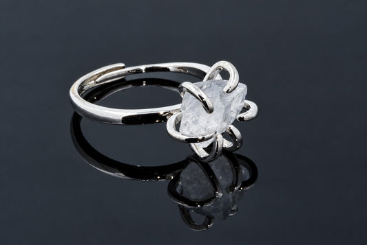 A silver-tone ring holds a rough clear crystal in curved arms on a dark reflective surface.