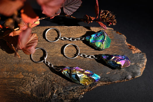 Keychains with rough blue, green and purple iridescent stones and silver-tone chains rest on a wooden surface beside red leaves.