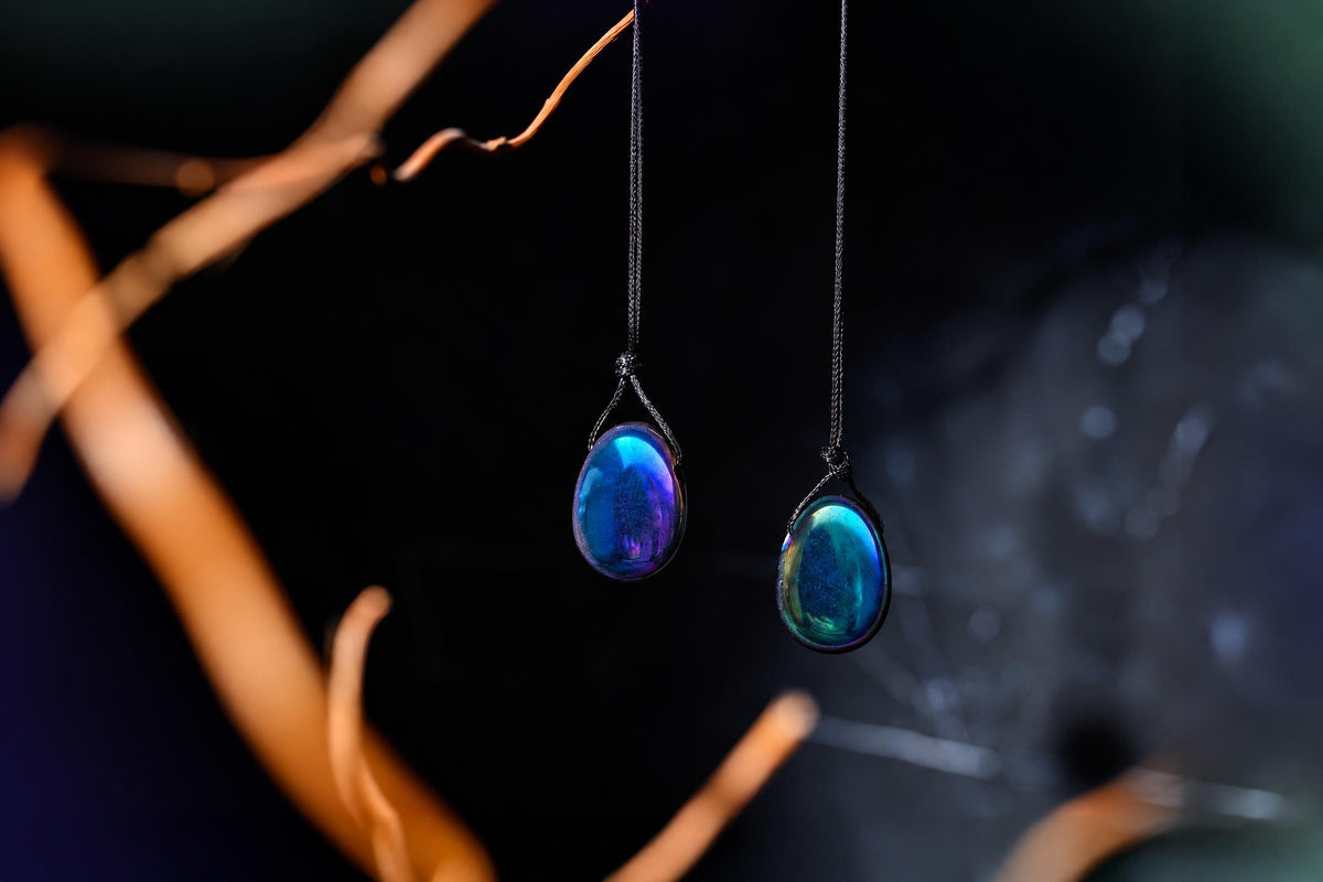 Two glossy blue, purple and green teardrop pendants hang from black cords among brown branches against a dark background.