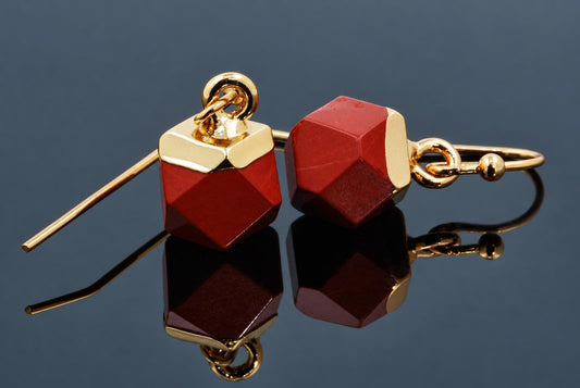 A pair of red faceted cube earrings with gold-coloured caps and hooks rests on a dark reflective surface.