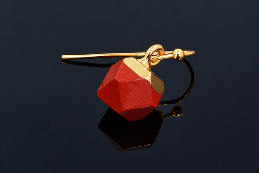 A gold-coloured ear clip earring with a red faceted cube pendant on a dark reflective surface.