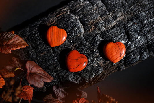 Polished red-orange heart-shaped stones with dark veins rest on a dark textured surface beside reddish leaves.