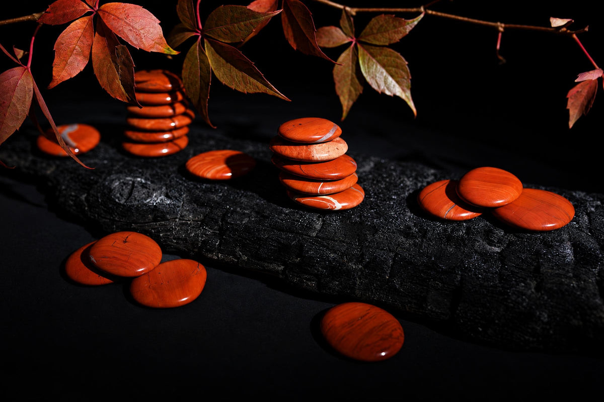 Polished red jasper palmstones are stacked and scattered on a dark textured surface beneath red leaves.