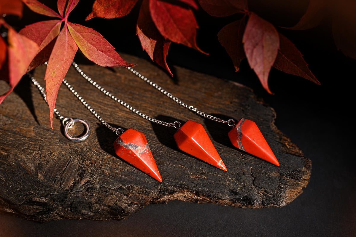 Red faceted pendulums on silver-tone chains lie on a wooden surface beneath reddish leaves.