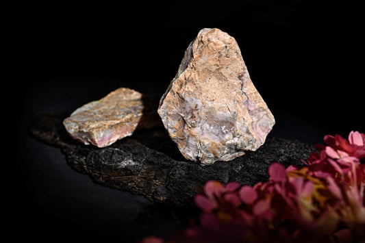 A rough pink-and-beige stone stands beside a flatter piece on a dark surface, with blurred pink flowers.