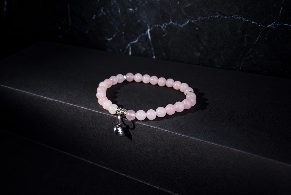 A pink round-bead bracelet with a dangling silver-tone heart charm rests on a dark surface with a veined backdrop.