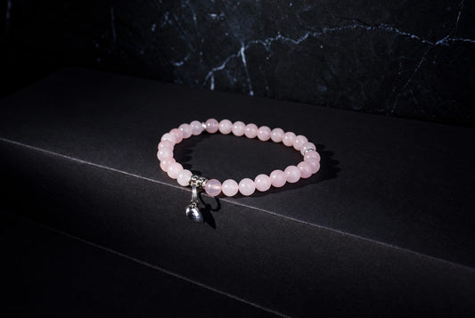 A pink round-bead bracelet with a dangling silver-tone heart charm rests on a dark surface with a veined backdrop.