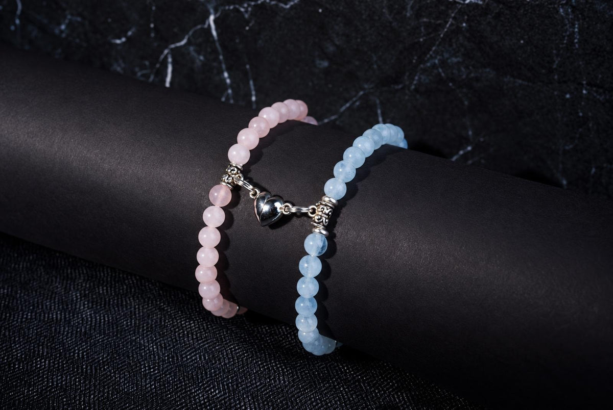 Two beaded bracelets on a black display stand, one pink and one light blue, connected by a silver heart charm.
