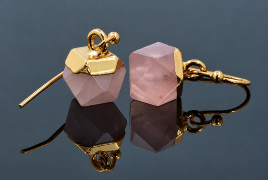 A pair of pale pink faceted cube earrings with gold-coloured caps and hooks rests on a dark reflective surface.