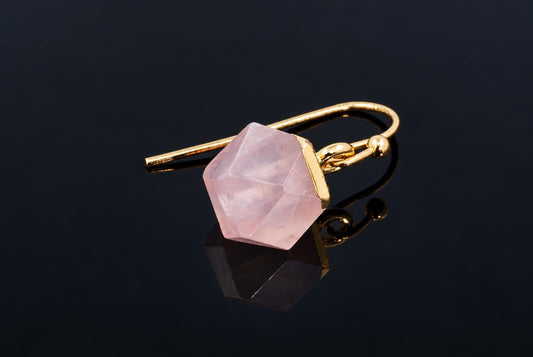 A pale pink faceted cube earring with a gold-coloured hook rests on a dark reflective surface.
