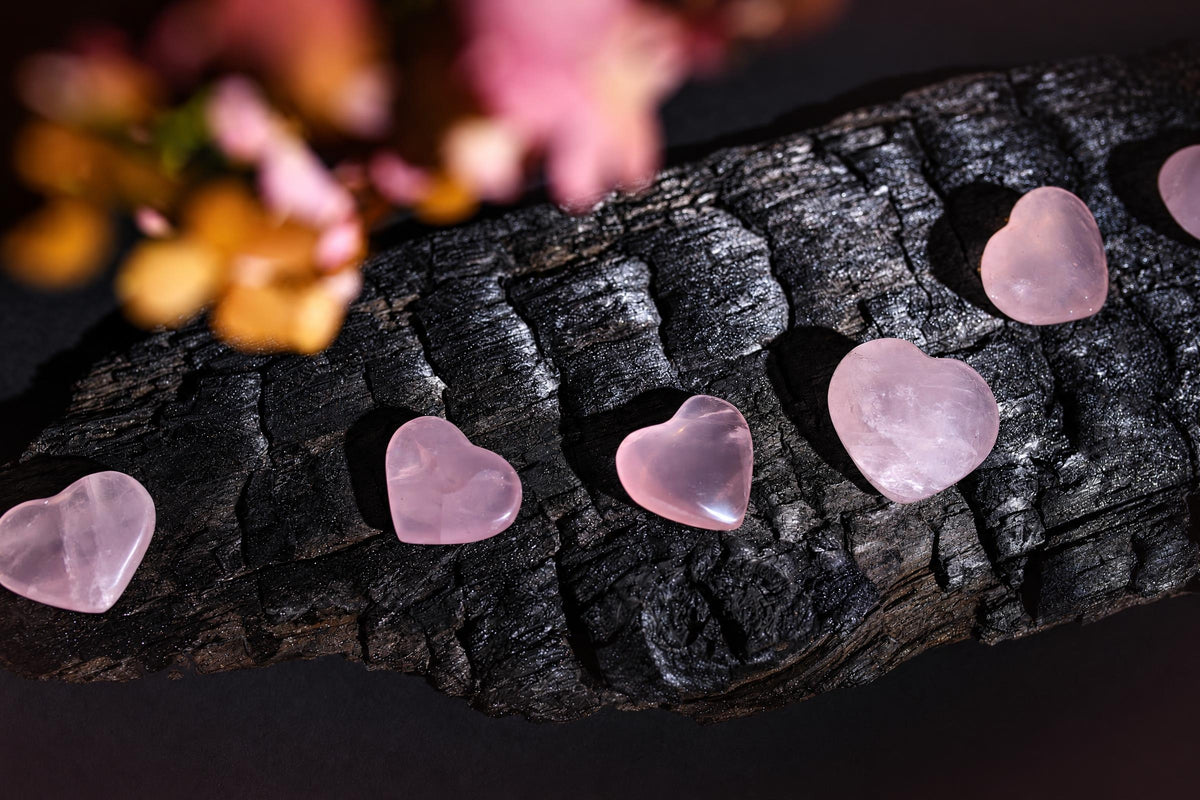 Polished pale pink heart-shaped stones form a row on a dark textured surface beneath blurred pink foliage.