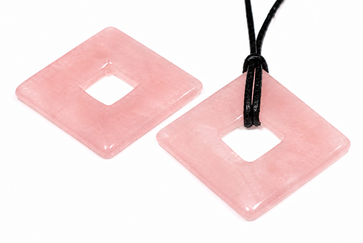 Two square, pink rose quartz pendants with a central hole lie on a white surface; one has a black cord.