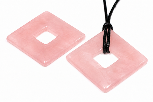 Two square, pink rose quartz pendants with a central hole lie on a white surface; one has a black cord.
