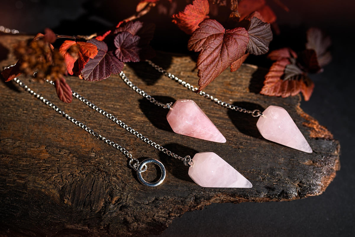 Pale pink faceted pendulums on silver-tone chains lie on a wooden surface beneath reddish leaves.