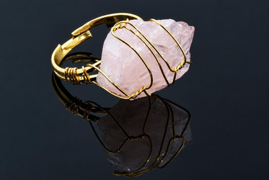 A gold-coloured ring holds a rough pale pink crystal wrapped in thin wire on a dark reflective surface.