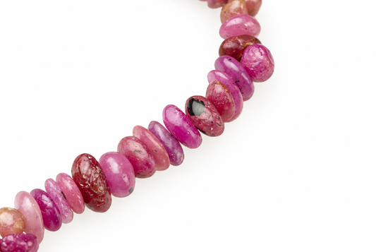 A string of polished, oval beads in shades of pink and red with dark spots on a white surface.
