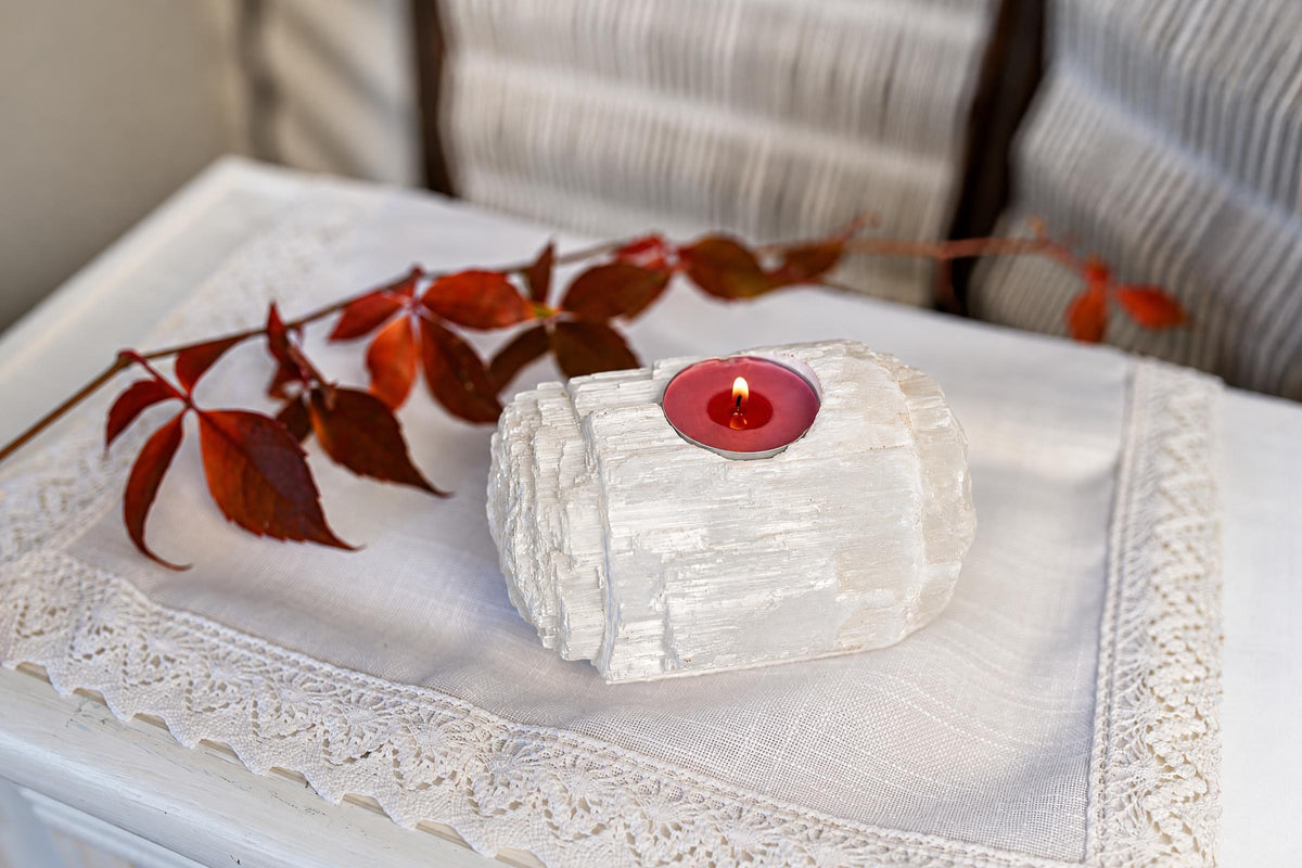 A red tealight burns in a ridged white crystal holder on pale lace-trimmed cloth beside reddish leaves.