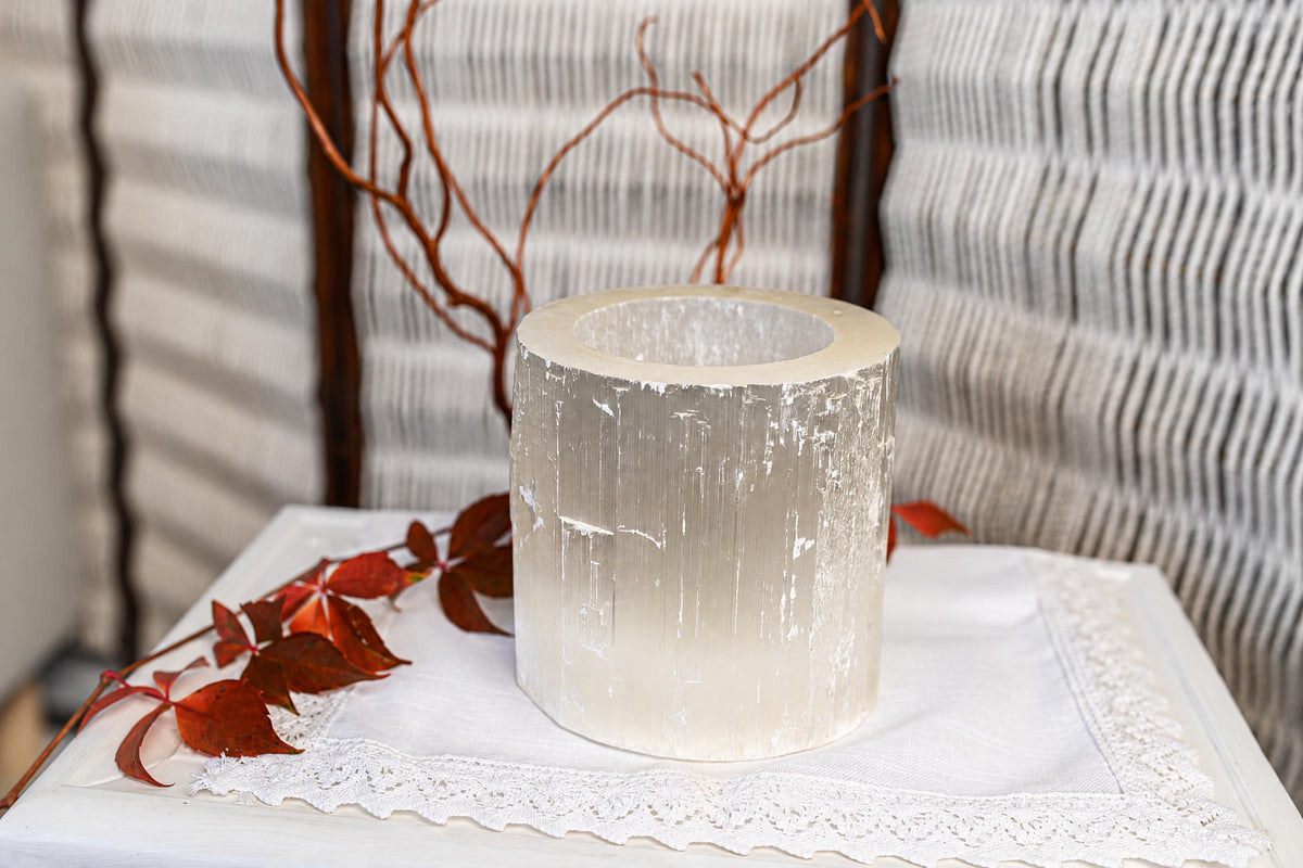 A white cylindrical candle holder with a hollow centre rests on pale cloth beside red leaves and thin brown branches.