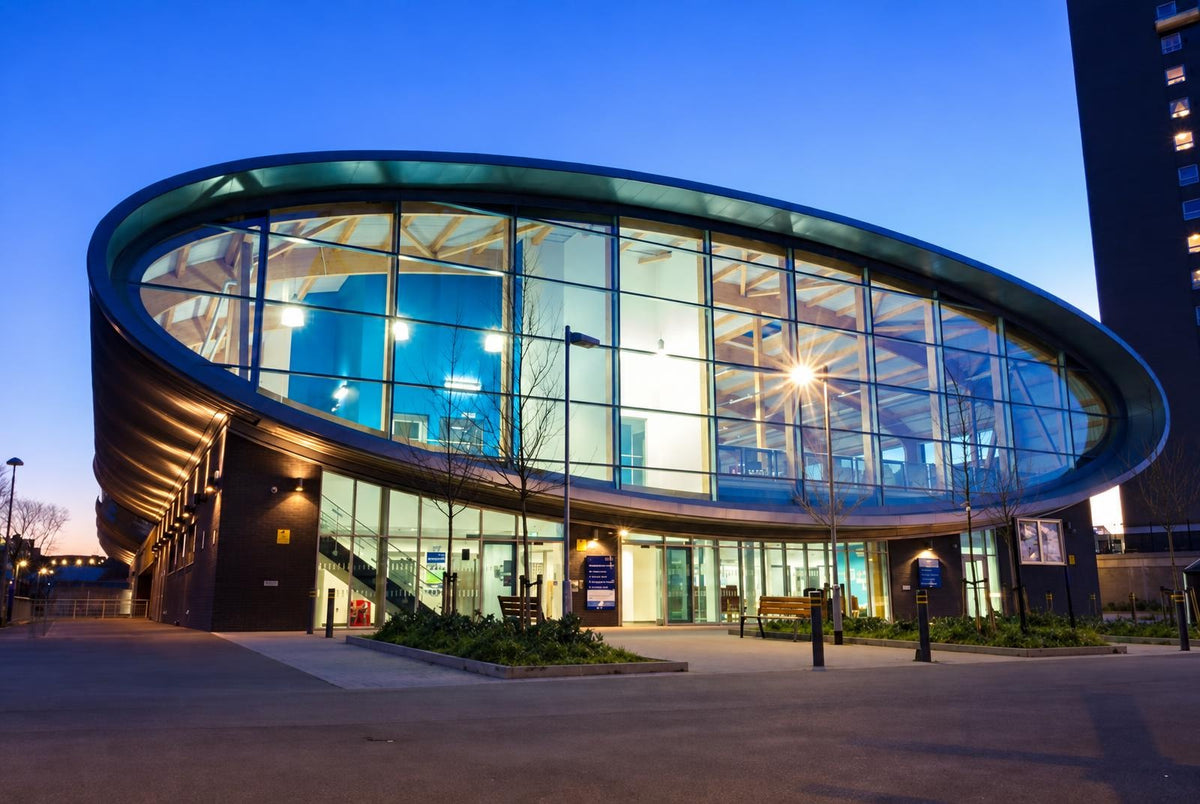 A modern building with a curved glass facade and dark brick base, illuminated at dusk against a blue sky.