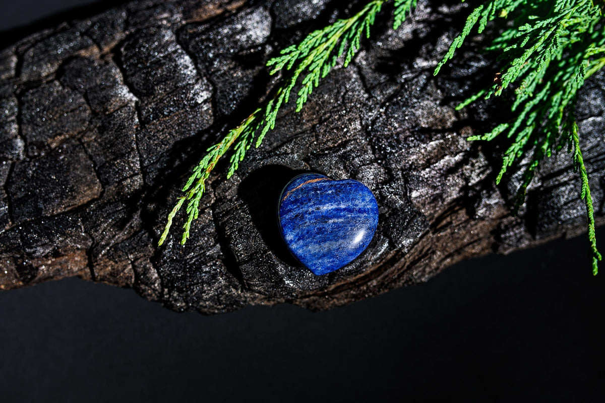 A glossy blue heart-shaped stone with pale streaks rests on a dark textured surface under green sprigs.