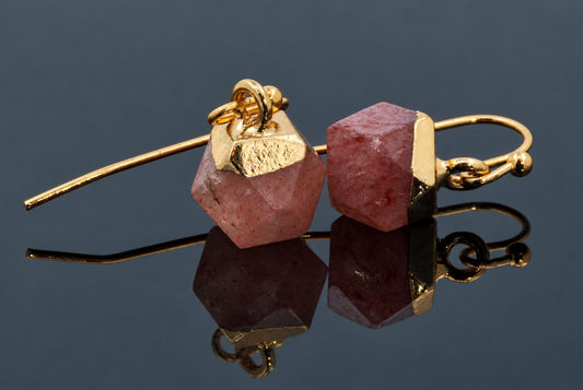 A pair of pink speckled faceted cube earrings with gold-coloured caps and hooks rests on a dark reflective surface.