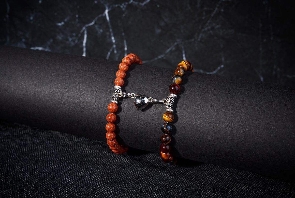 Orange and striped brown-and-gold beaded bracelets joined by a silver-tone heart charm on a black display.