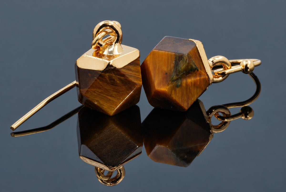 A pair of brown-and-gold faceted cube earrings with gold-coloured hooks rests on a dark reflective surface.
