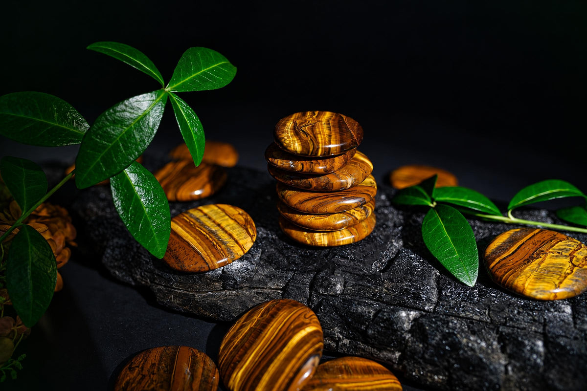 Polished golden-brown oval stones with dark bands are stacked and scattered on a dark surface among green leaves.