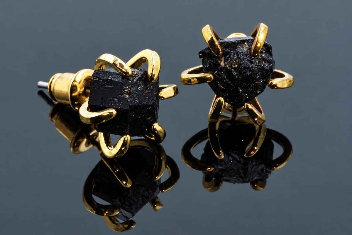 Rough black crystal stud earrings in curved gold-coloured settings rest on a dark reflective surface.
