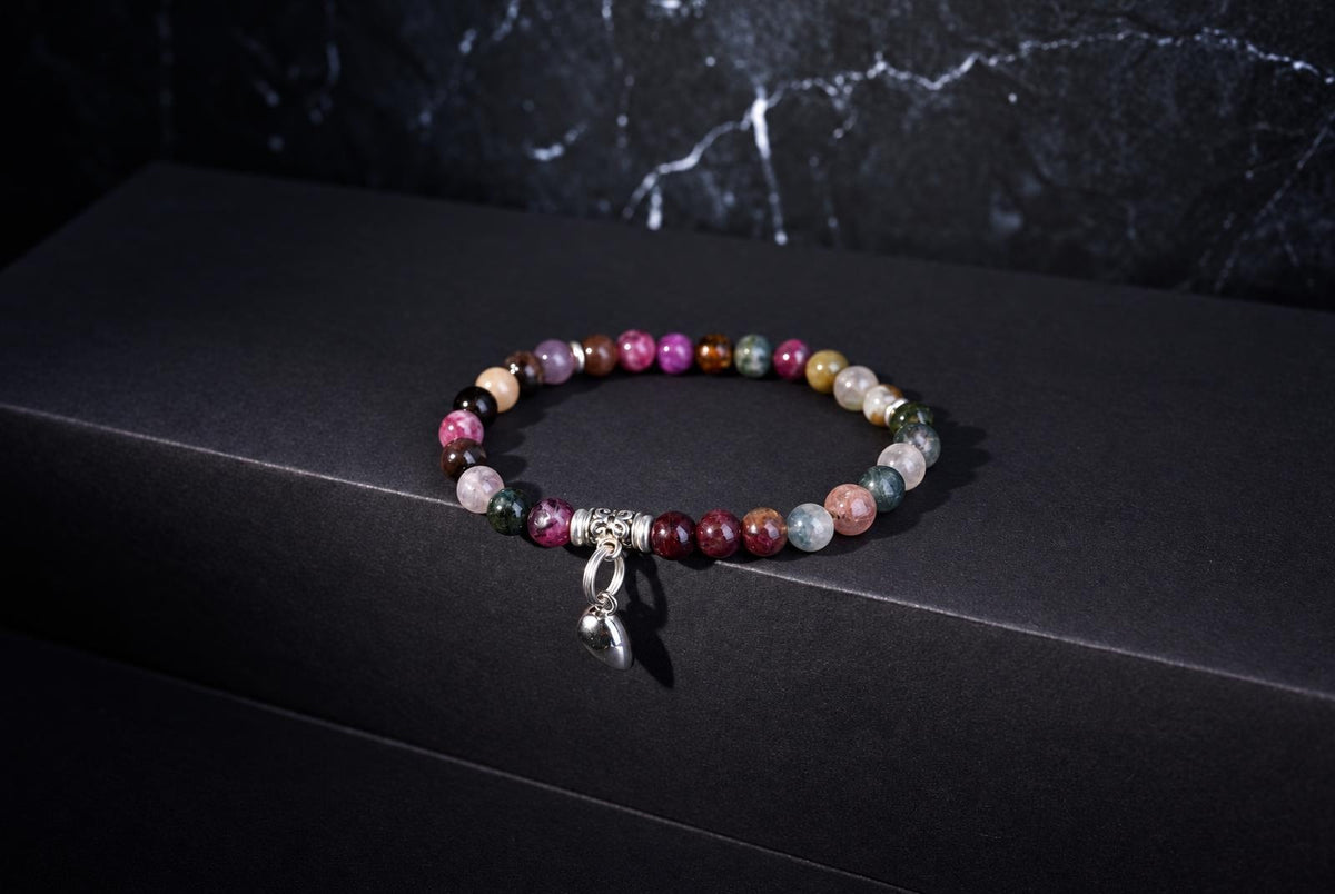 A multicoloured beaded bracelet with a dangling silver-tone heart charm on a dark surface and veined backdrop.