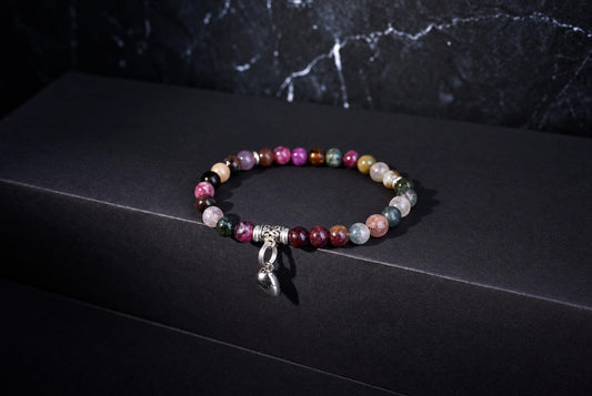A multicoloured beaded bracelet with a dangling silver-tone heart charm on a dark surface and veined backdrop.